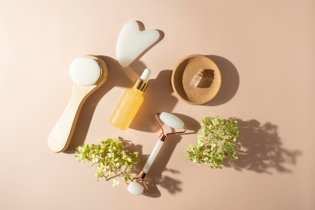 A face roller and quartz massager in the form of the heart a face brush and a face serum with vitamin C lying on the beige background