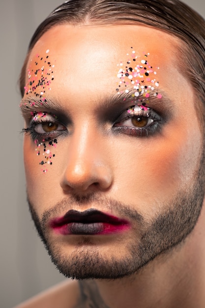 Face of sad young bearded man with colorful glitters on face and black lips, musical actor concept