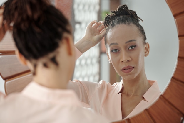 Face of serious attractive young woman making hairbun and looking at mirror in the morning