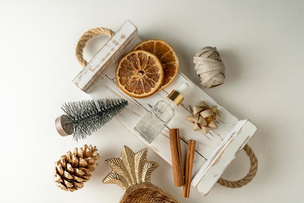 A face serum or essential oil for Christmas holidays in a red dropper bottle lying on a white background with golden bows cinnamon and dried oranges around