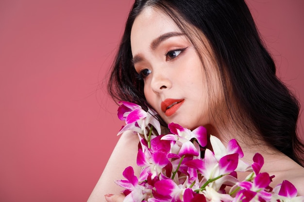 Face shot Portrait of Fashion 20s Asian Woman beautiful black hair express feeling sensual happy smile. Girl hold purple orchid flower for skin care cosmetic and treatment over pink Background