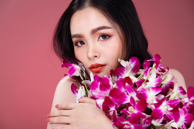 Face shot Portrait of Fashion 20s Asian Woman beautiful black hair express feeling sensual happy smile. Girl hold purple orchid flower for skin care cosmetic and treatment over pink Background