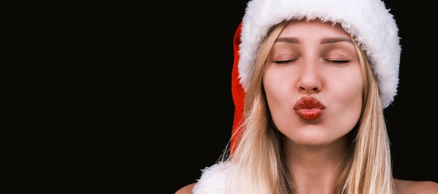 Face shot of young attractive blonde woman with plump lips in christmas hat sends air kiss against d