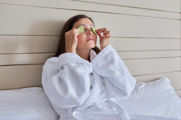 Face skin care of middle aged woman. Mature female in bathrobe doing home spa treatments with cucumbers, natural organic cosmetics and anti-aging facial massage at home in bed