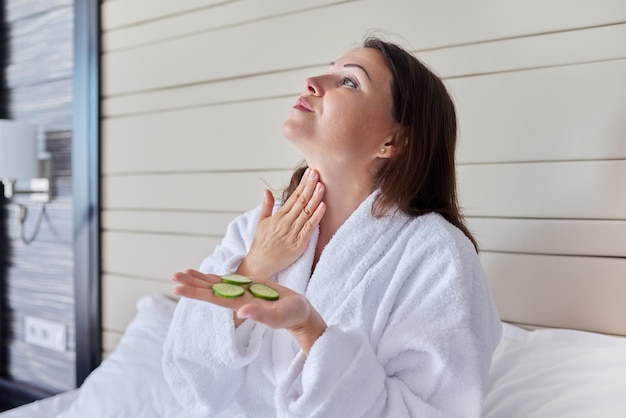 Face skin care of middle aged woman. Mature female in bathrobe doing home spa treatments with cucumbers, natural organic cosmetics and anti-aging facial massage at home in bed