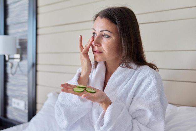 Face skin care of middle aged woman. Mature female in bathrobe doing home spa treatments with cucumbers, natural organic cosmetics and anti-aging facial massage at home in bed