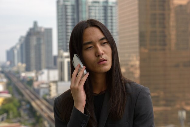 Face of young Asian businessman with long hair using phone in the city outdoors