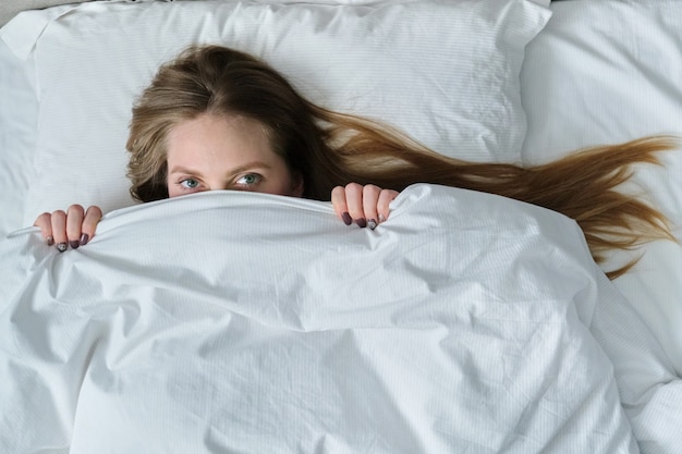 Face of young beautiful woman lying on pillow in bed with blanket, posing girl smiling covering her face