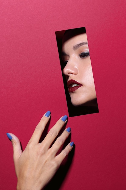 Foto volto di giovane bella donna con un trucco luminoso e labbra rosse guarda attraverso un buco in carta rossa.