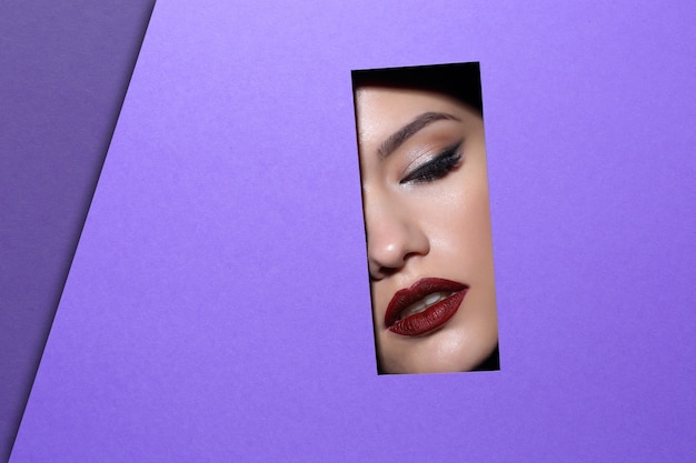 Face of young beautiful woman with a bright make-up and red lips looks through a hole in violet paper. 