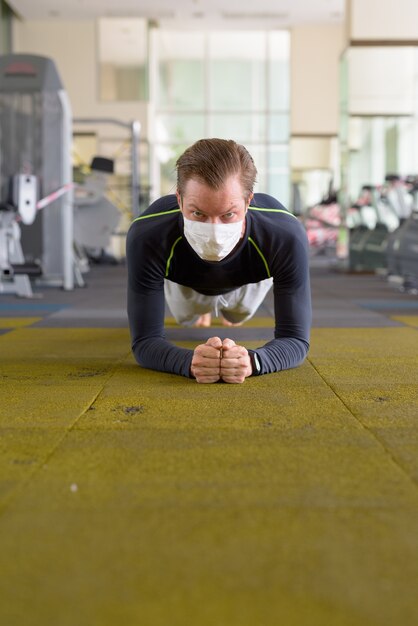 Face of young man with mask doing plank position on the floor at gym during coronavirus covid-19