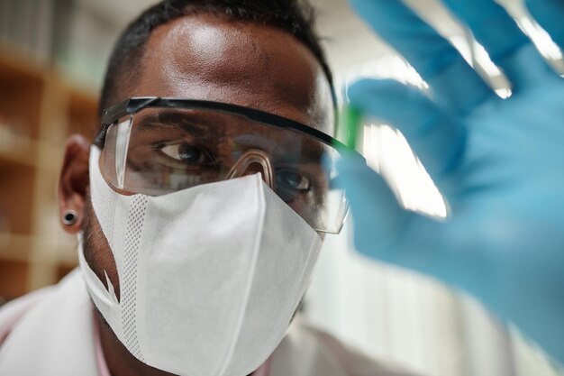 Face of young pharmacist in protective eyeglasses and mask looking at pill in his gloved hand while