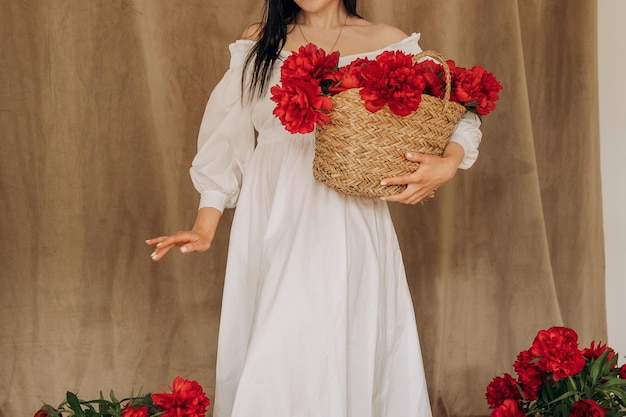 A faceless brunette girl in a white delicate dress holds a basket with red peonies in her hand Spring flowers for a gift