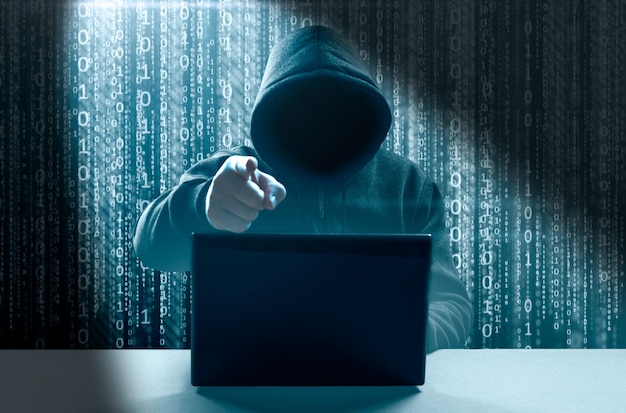 Faceless hooded hacker showing index finger gesture hacker man\
with laptop attack to server network system online in data internet\
security hacking concept dark binary background