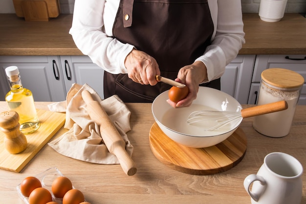 A faceless hostess in a brown apron breaks a raw egg with a knife into a bowl of milk to make dough ingredients for cooking on the countertop