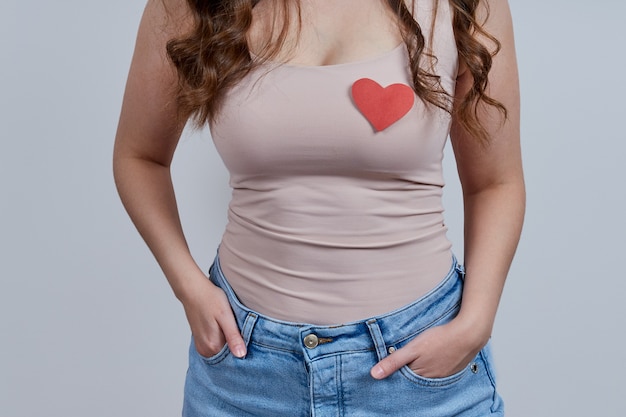 A faceless image of a woman in a pink T-shirt, with a heart symbol on her chest. Studio shot over gray background.