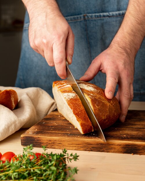 Faceless man cutting fresh home baked crusty bread with large knife on wooden Board on kitchen table. Homemade bakery, vertical
