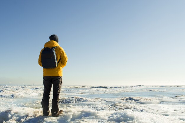 Faceless man in yellow raincoat with backpack standing and looking on frozen sea