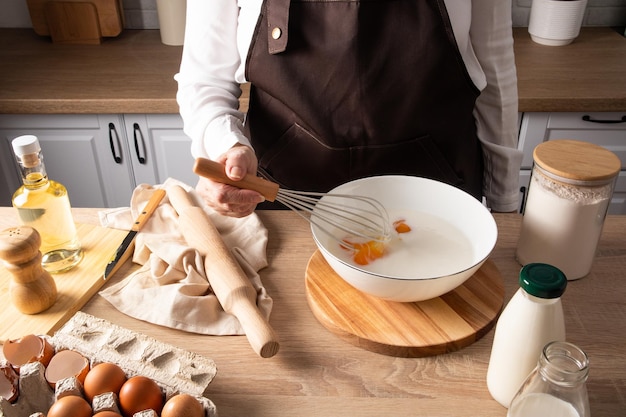 A faceless middleaged woman in an apron whisks an egg with milk in a bowl to make the dough can of flour a bottle of milk on the table