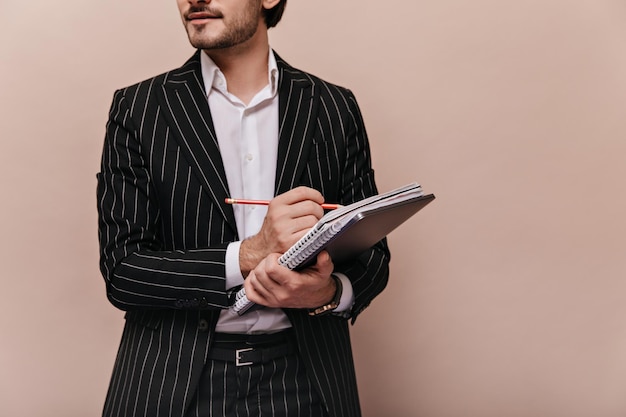 Faceless photo of man in white shirt and black striped suit turning head aside while making notes and holding papers folders against beige background