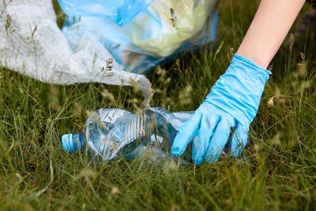 Faceless portrait of person hands picking up plastic bottle from ground, cleaning meadow and putting it to garbage bag, trash on grass, women hand in blue glove.