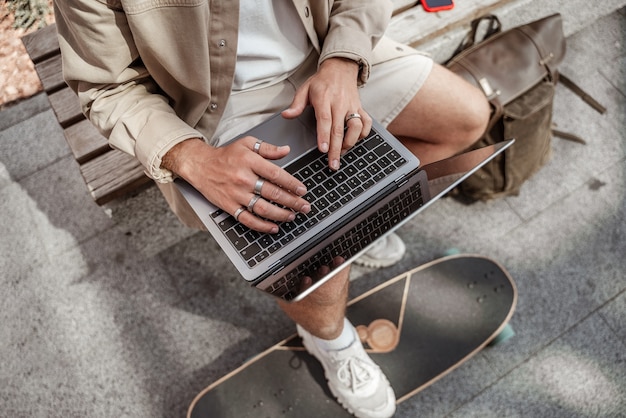 Faceless portrait of young man skater sitting in the street working on laptop with mobile phone with longboard and backpack. Remote working concept. Millennial hipster guy. Hands typing from above