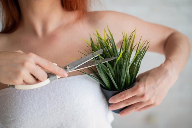 Faceless redhaired Caucasian woman wrapped in a white terry towel shears a potted plant with scissors Armpit hair removal concept Selfdepilation procedure Trim off excess vegetation