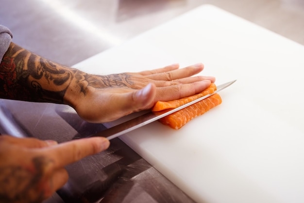 Photo faceless shot of tattooed chef cutting salmon in restaurant kitchen