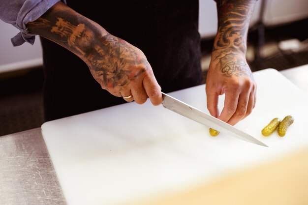 Faceless view of tattooed cook cutting marinated cucumbers on board in cafe