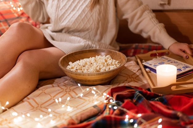 Faceless woman in knitted wool cozy sweater in bed on a blanket at camping mobile home trailer with a book and popcorn food and candle relaxing and enjoyment on happy new year and merry christmas eve