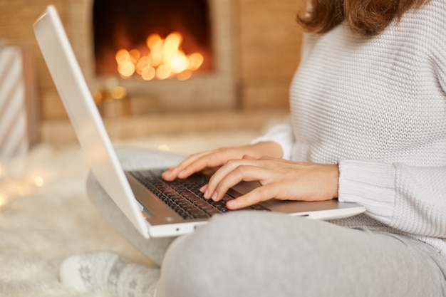 익명의 여성이 그녀의 노트북 컴퓨터에 메시지를 입력하고 그녀는 뜨거운 불이 타는 곳 근처에서 이완되고, 알 수없는 여성이 다리를 건너 바닥에 앉아 그녀의 무릎에 hotebook을 들고 온라인으로 작업합니다.