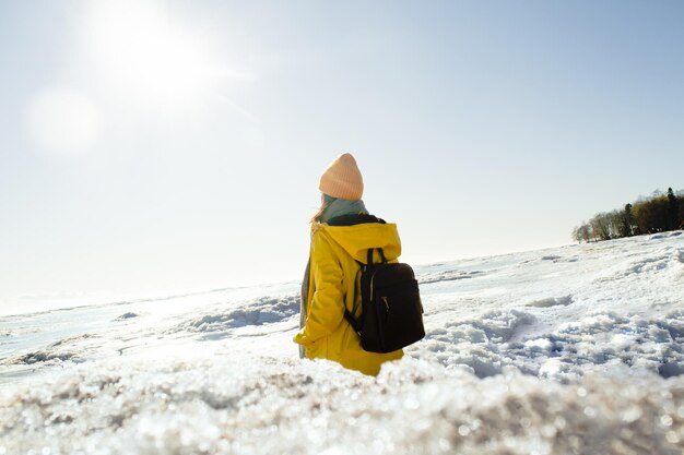 Foto donna senza volto in impermeabile giallo con zaino in piedi e guardando il mare ghiacciato