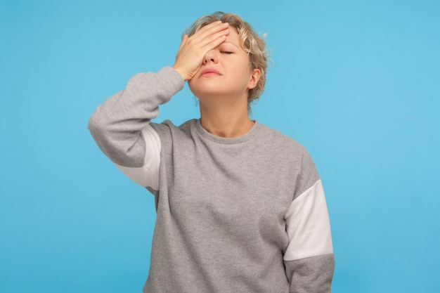 Facepalm Hopeless desperate woman in casual grey sweatshirt covering face with hand feeling shame sorrow regret for mistake worried about failure indoor studio shot isolated on blue background