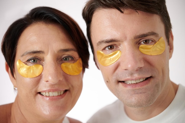 Faces of smiling mature man and woman wearing hydrogel patches skincare and beauty concept