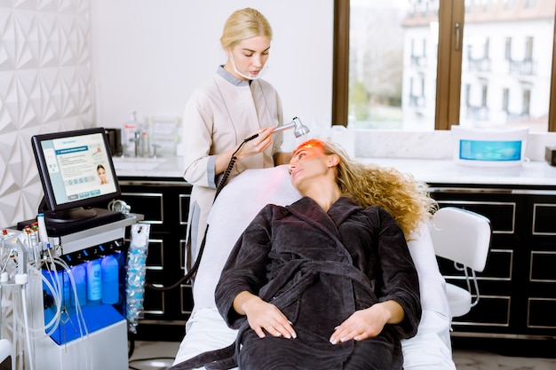 Facial beauty treatment in modern cometology clinic. Pretty blond curly woman having red led light therapy