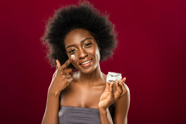 Facial cream. Positive young woman using organic cream while standing against red wall