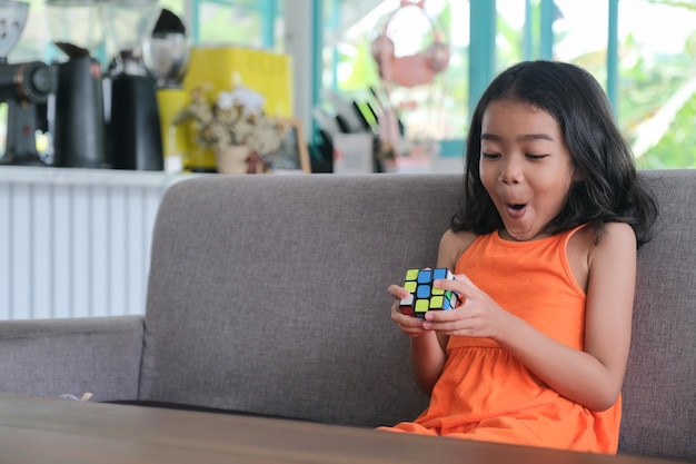 Photo facial expression of little girl sitting alone in the sofa playing rubik's cubes