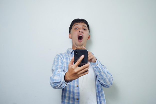 Выражение лица очень шокированного азиатского мужчины с помощью мобильного телефона на белом фоне