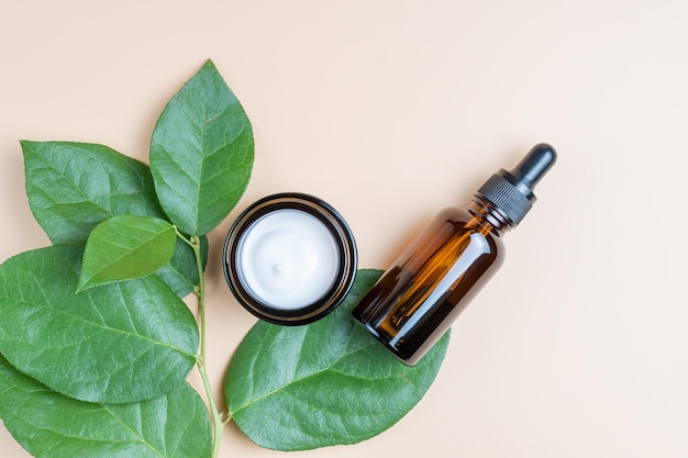 Facial serum dropper and moisturizer cream in brown glass bottle and jar on fresh green tropical leaves background Eco organic natural beauty skincare products top view mockup for cosmetic brand