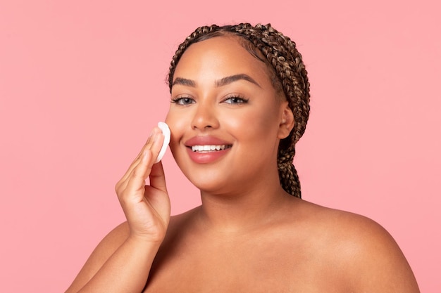 Facial skincare concept Happy black chubby woman using cotton pad caring for smooth skin posing over pink background