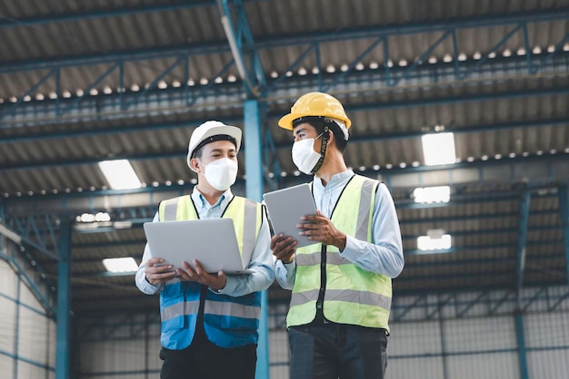 Factory manager and engineer in  medical mask and safety protective suite using computer laptop and digital tablet working discuss together