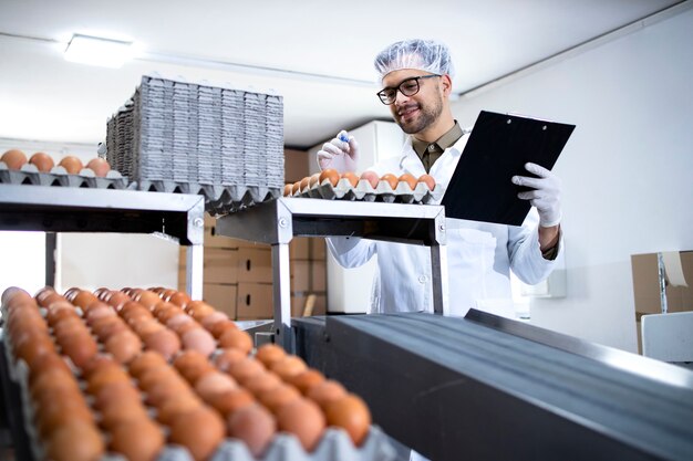 Factory worker holding checklist inspecting and checking quality of chicken eggs at food processing plant.