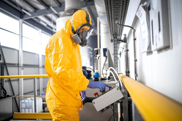 Factory worker in protection suit and gas mask operating machine inside chemicals plant