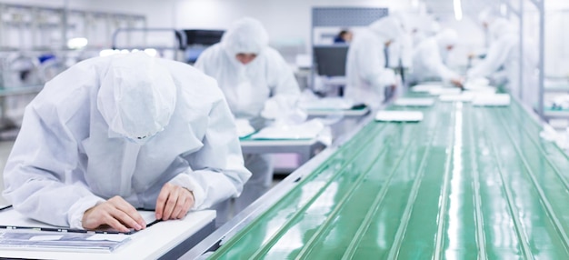 Foto lavoratori di fabbrica in tute bianche da laboratorio e maschere facciali che producono televisori su una linea di montaggio verde sfondo sfocato
