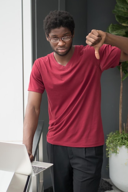 Photo failed college student pointing thumb down; portrait of unhappy, angry, upset nerd man, college student pointing thumb down, bad, no, rejecting concept; african young adult man model