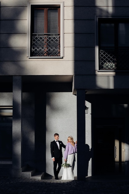 Fair-haired bride wearing wedding dress and lilac jacket with bouquet in her hand and bridegroom in black suit and white shirt standing close to each other at contrasting grey wall outdoors