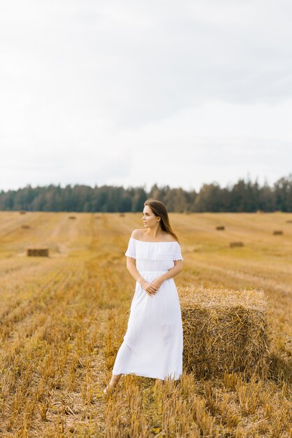 fair haired young woman farmer in a white dress in a field with stacks of straw enjoys a summer day