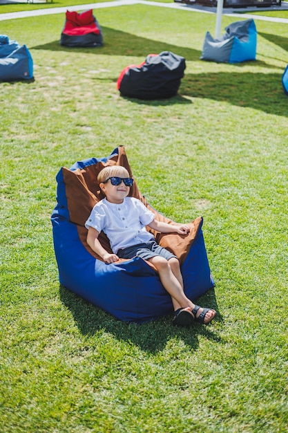 A fairhaired boy of 56 years old in a white Tshirt and shorts is sitting on a bean bag chair A child in sunglasses rests in the fresh air