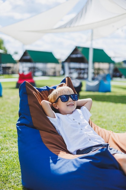 A fairhaired boy of 56 years old in a white Tshirt and shorts is sitting on a bean bag chair A child in sunglasses rests in the fresh air