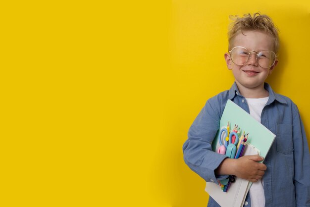 A fairhaired boy holding school supplies and grimaces on a yellow background copy space